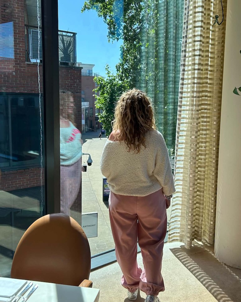 Jo Cox standing by a tall window in a bright restaurant, looking out at the street